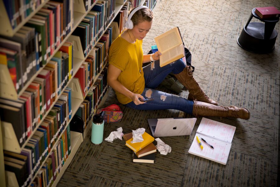 Student studying on the floor of the library sitting against a bookshelf.