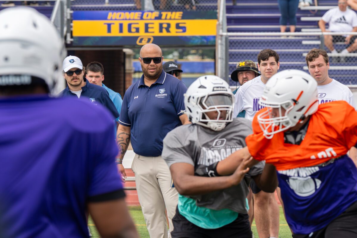 Coaches from St. Ambrose University and St. Norbert College observe linemen battling during a drill at the 2025 Avante Mitchell Football Mega Camp at Olivet Nazarene University. "Home of the Tigers" sign is visible in the background.

