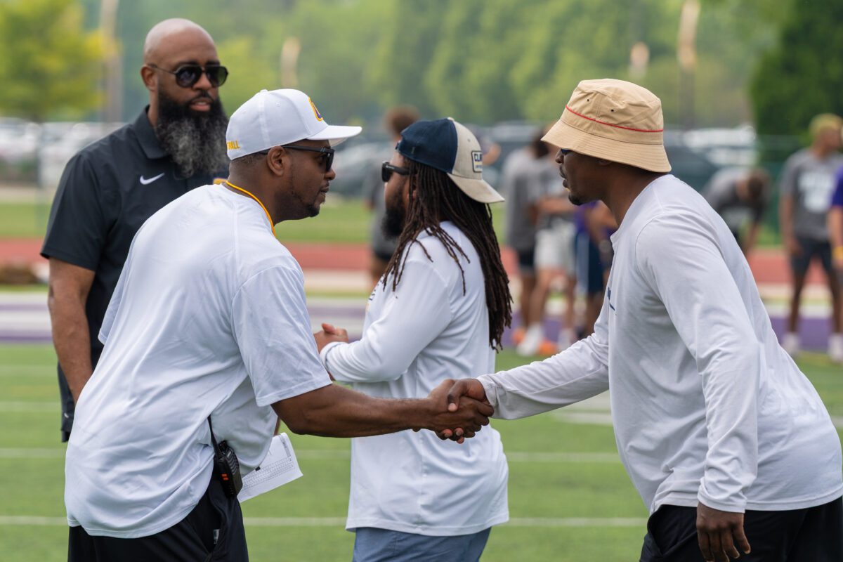 Coaches shake hands on the field at the 2025 Avante Mitchell Football Mega Camp, hosted at Olivet Nazarene University. One coach wears a white Olivet shirt, while another sports a tan bucket hat. Other staff and players are visible in the background.

