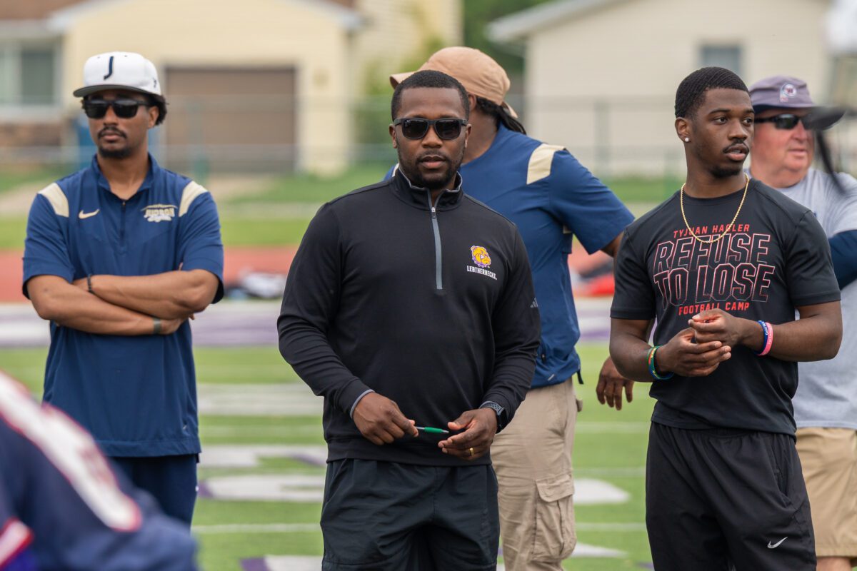Coaches from Judson University and Western Illinois University observe athletes during a drill at the Avante Mitchell Football Mega Camp. The coach in black sports a Leathernecks logo, while another wears a "Refuse to Lose Football Camp" t-shirt.