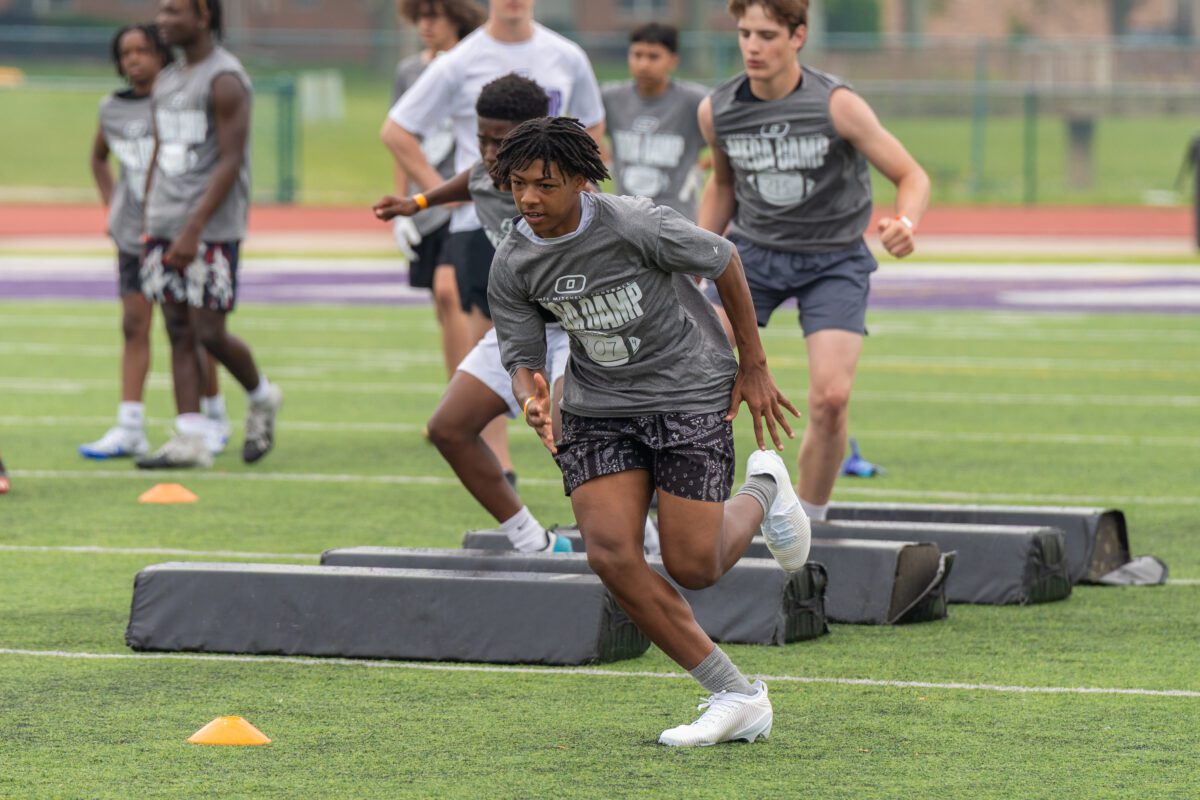 High school football players sprint through agility drills during a college recruiting camp on a turf field.

