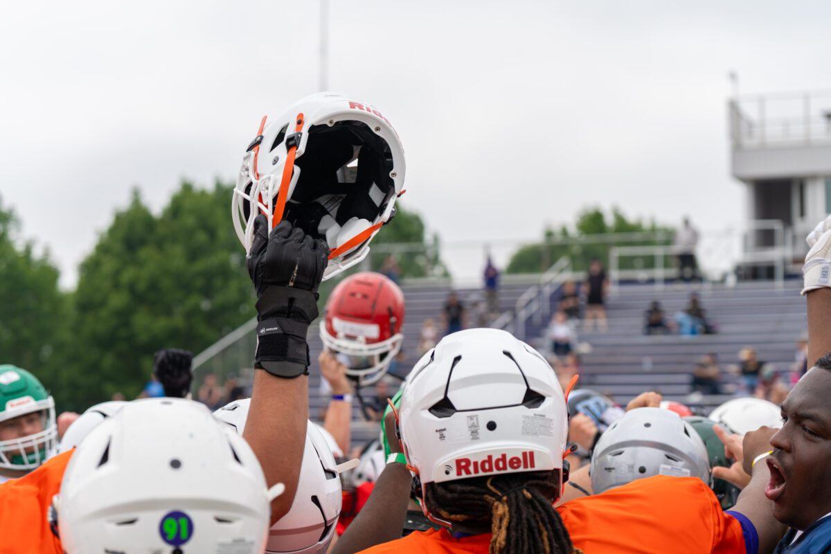 A group of high school football players raise their helmets in the air during a team huddle at the 2025 Avante Mitchell Football Mega Camp, held at Olivet Nazarene University. Players wear various helmet colors and practice jerseys as they unite on the field.

