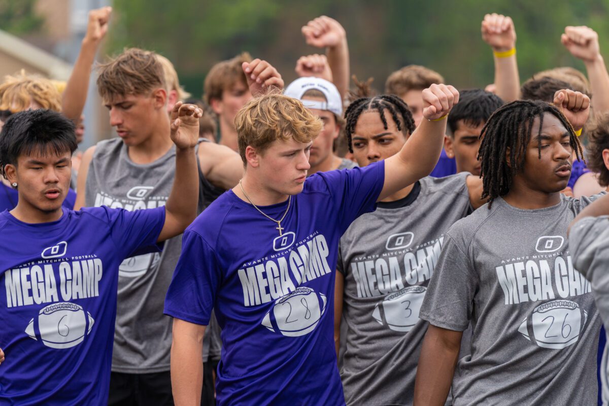 Coaches from St. Ambrose University and St. Norbert College observe linemen battling during a drill at the 2025 Avante Mitchell Football Mega Camp at Olivet Nazarene University. "Home of the Tigers" sign is visible in the background.

