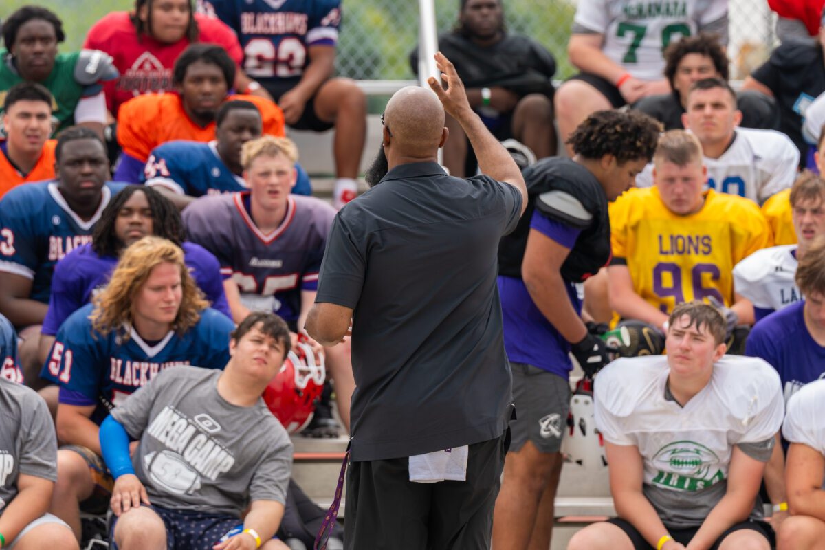 A coach addresses a large group of high school football players seated on bleachers during the Avante Mitchell Football Mega Camp. Players wear a mix of jerseys representing different teams, including Blackhawks, Lions, and Irish.