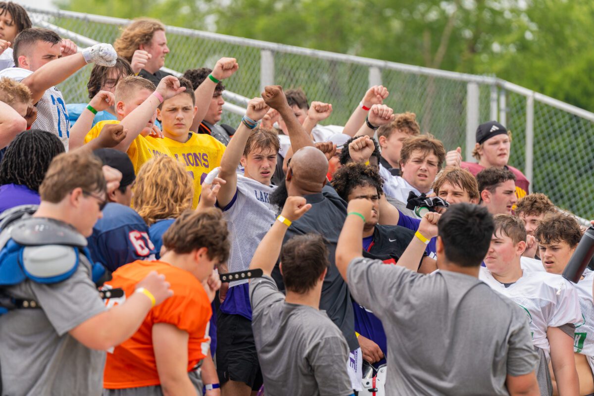 A group of high school football players raises their fists in a unified cheer around a coach during the Avante Mitchell Mega Camp, standing on metal bleachers with shoulder pads and jerseys from various schools.


