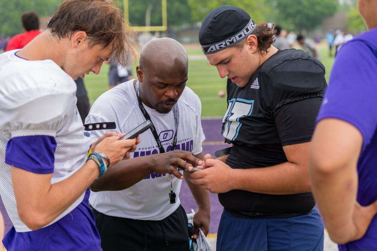 A coach wearing an "Avante Mitchell Football Mega Camp" shirt closely reviews a phone screen with two high school football players, one in black gear and the other in white and purple, during a break on the field.