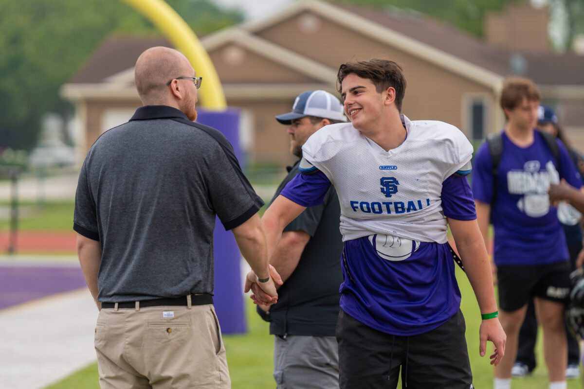 A football coach from the University of Saint Francis, wearing a gray "USF Football" hat and navy jacket, stands next to a student-athlete in a purple "Mega Camp" shirt as they both look down at their phones during a football showcase.

