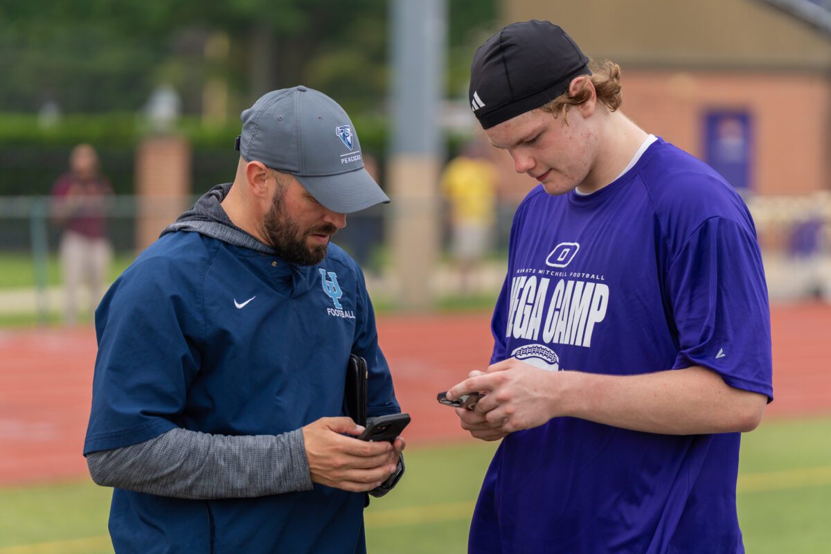 A football coach from the University of Saint Francis, wearing a gray "USF Football" hat and navy jacket, stands next to a student-athlete in a purple "Mega Camp" shirt as they both look down at their phones during a football showcase.


