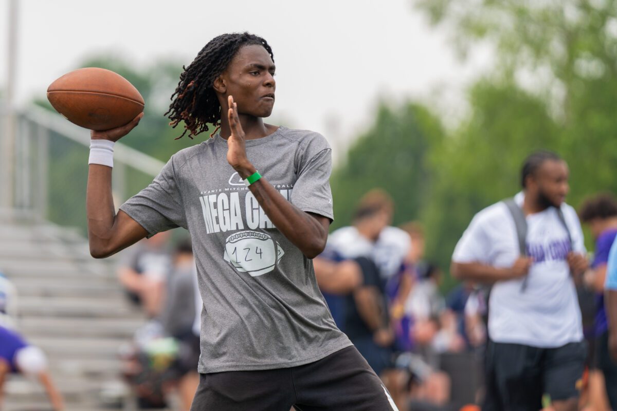 A high school football player wearing a gray "Mega Camp" shirt and green gloves reaches out to catch a football mid-air during a drill on a turf field.

