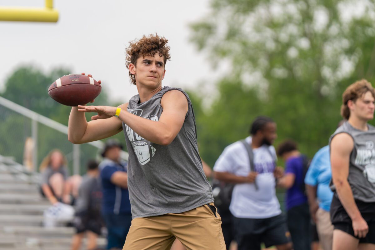 A young quarterback in a gray sleeveless “Mega Camp” shirt winds up to throw a football during a drill, with other athletes and coaches blurred in the background on a football field.

