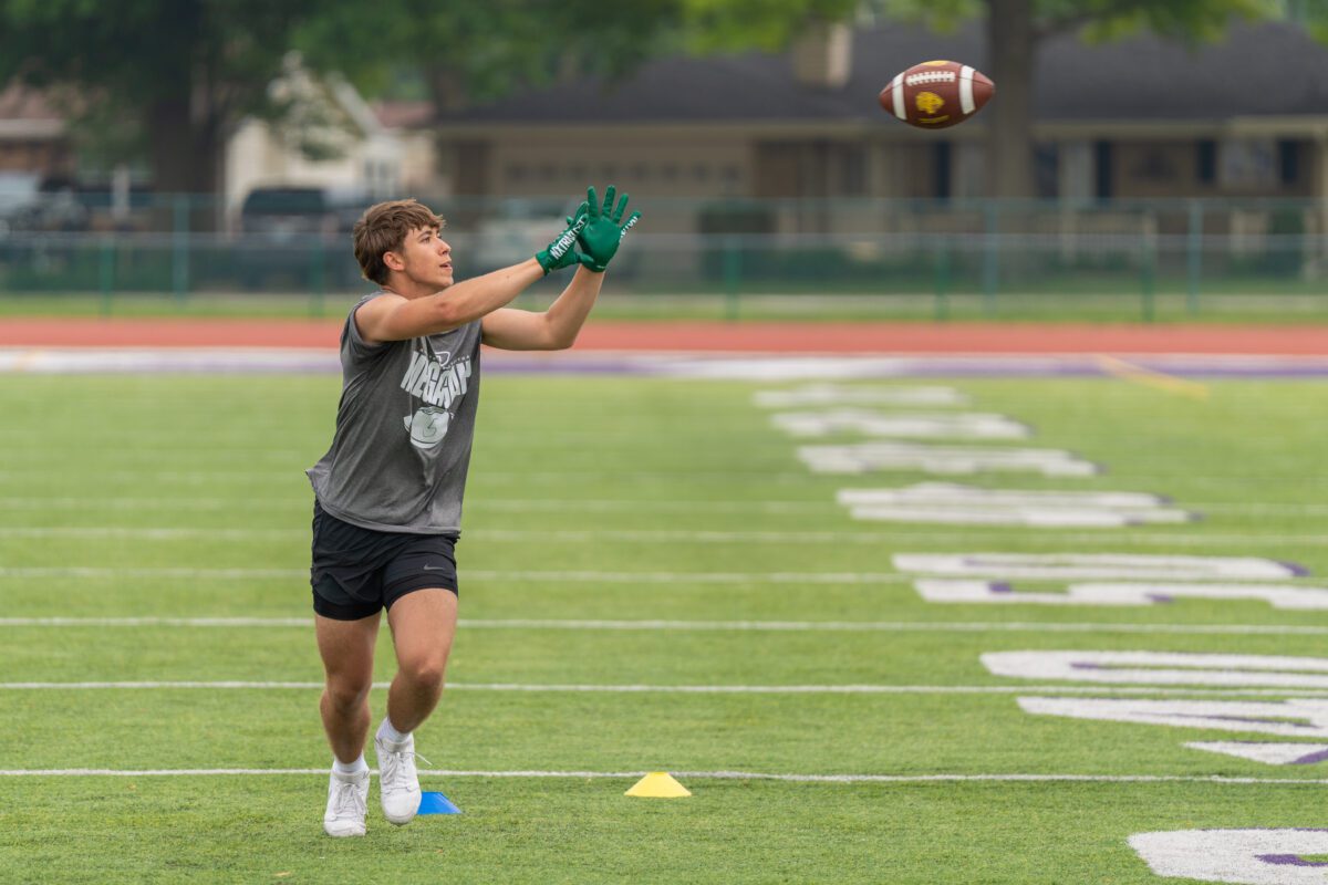 A high school football player wearing a gray "Mega Camp" shirt and green gloves reaches out to catch a football mid-air during a drill on a turf field.
