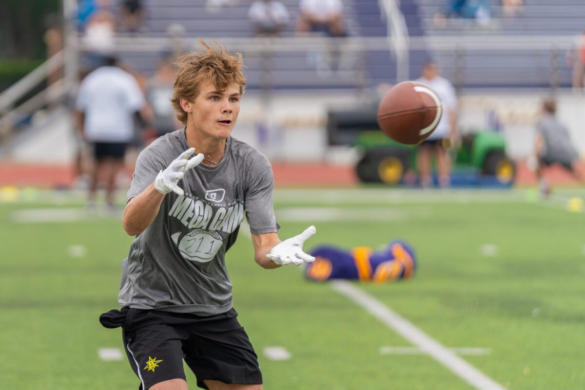 A focused young athlete at Mega Camp reaches forward with gloved hands to catch a football mid-air during a drill, wearing a gray event shirt and black shorts on a green turf field.