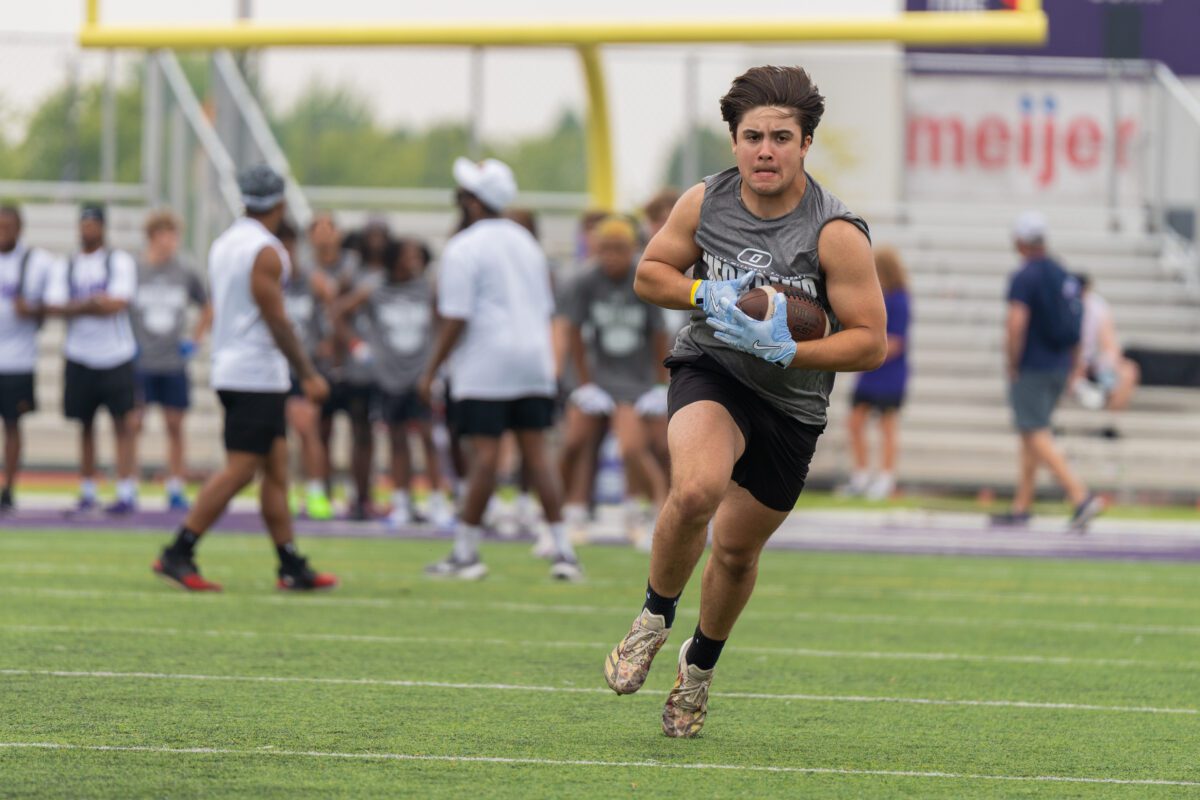 A determined high school football player in a gray “Mega Camp” shirt and light blue gloves sprints forward while gripping a football tightly during a drill, with teammates and coaches watching in the background.
