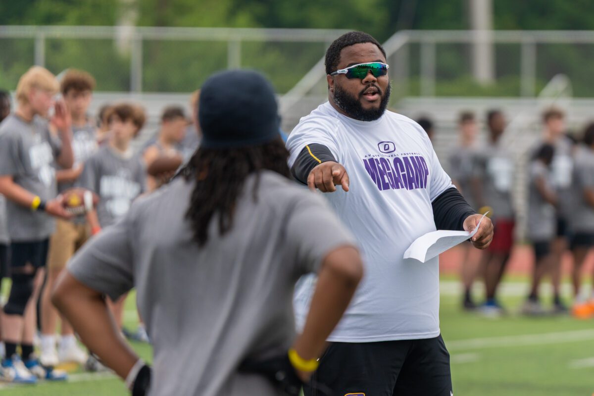 Khalid Hill, former University of Michigan fullback and Seattle Seahawk, gestures while coaching at the Avante Mitchell Football Mega Camp, wearing a white “Mega Camp” shirt and reflective sunglasses on a sunny day
