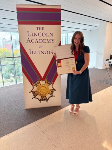 Grace Amburgey standing next to a Lincoln Academy of Illinois banner holding her award