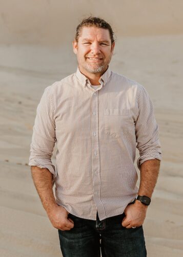 Steven standing in a desert looking at the camera. He is wearing a white and orange stripped long sleeve shirt rolled halfway up his arms.  