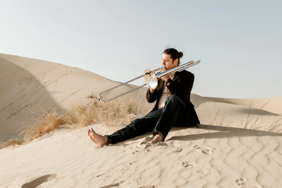 Steven sitting on a sand dune playing a trombone.
