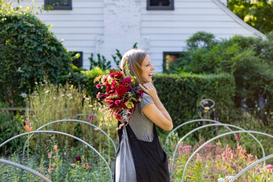 Jeanie holding flowers in a garden with a white house in the background.