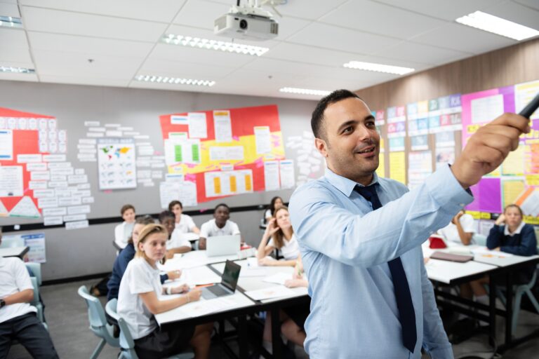 Classroom with diverse students and a male teacher pointing at board. Students engaged, teacher explaining. Classroom setting, teacher, students, learning. Teacher teaching a diverse high school class